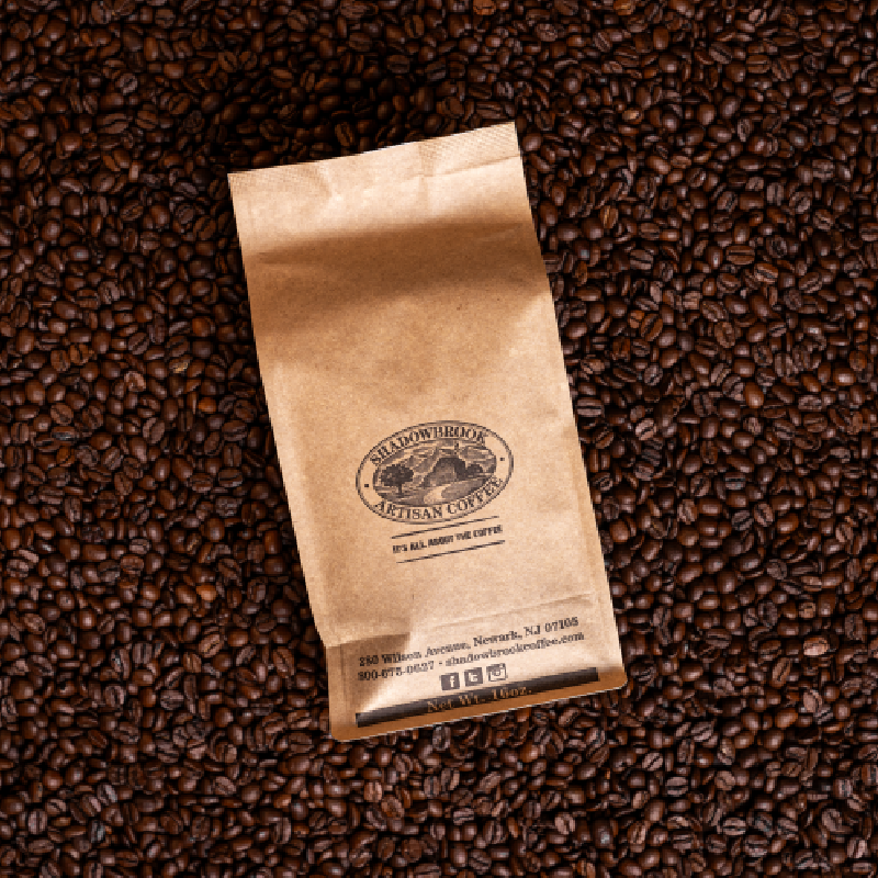A 16oz bag of Shadowbrook Artisan Coffee sitting on a bed of coffee beans. 