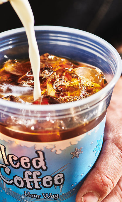Milk pouring into a plastic iced coffee cup filled with iced coffee, being held by a hand .