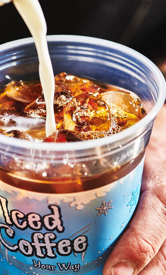 Milk pouring into a plastic iced coffee cup filled with iced coffee, being held by a hand .